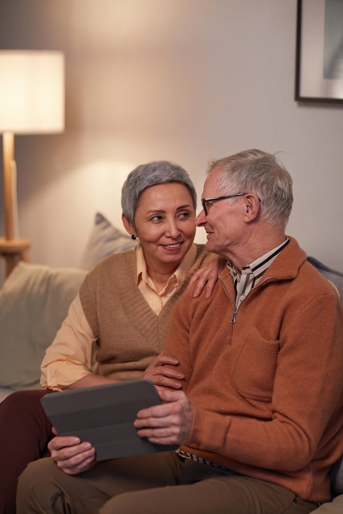 Happy elderly couple sitting on sofa, using tablet, feeling love and joy.
