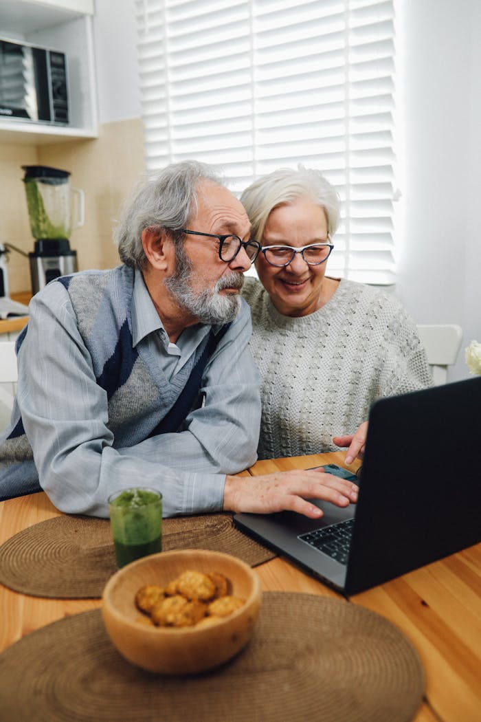 Elderly couple enjoying time together on a laptop in their cozy home kitchen.